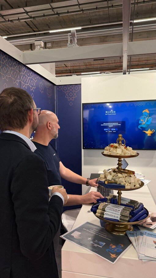 Bildbeschreibung: Zwei Männer vor einem Bildschirm auf einer IT-Messe. Der Stand der IT-Agentur präsentiert innovative Softwarelösungen. Auf einem Tisch im Vordergrund befinden sich Snacks und Informationsmaterialien. Moderne, professionelle Atmosphäre.

