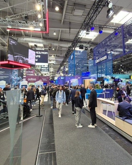 Menschen besuchen eine belebte Messehalle mit verschiedenen Technologie-Ausstellungsständen und digitalen Displays.