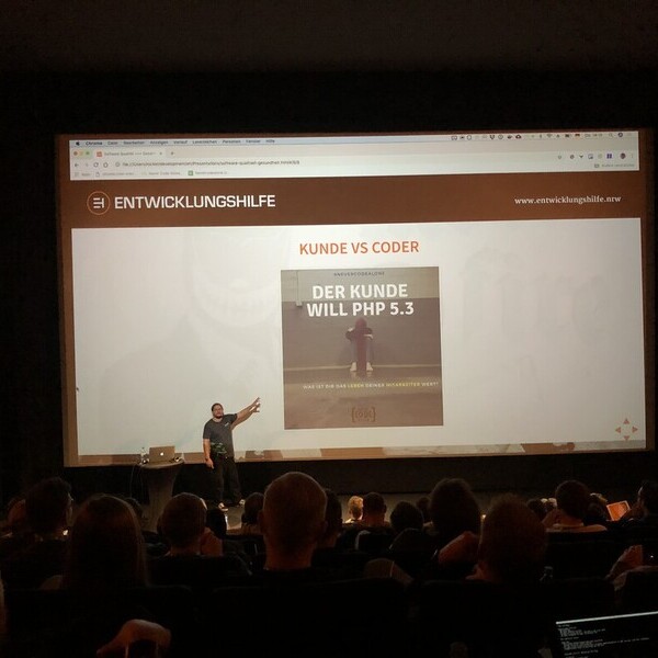 Titel: Vortrag über PHP bei IT-Entwicklungsagentur

Beschreibung: In einem professionellen Vortragsraum erklärt ein Referent eindrucksvoll das Thema „Kunde vs. Coder“ im Kontext von PHP-Versionen. Die Präsentation mit dem Thema „Der Kunde will PHP 5.3“ bi