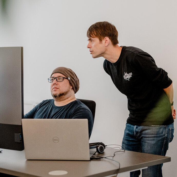 **Bildbeschreibung: IT-Experten bei der Zusammenarbeit im Büro**

In einem modernen IT-Büro diskutieren zwei Experten konzentriert über die neuesten technologischen Trends. Ein Entwickler sitzt vor mehreren Bildschirmen, während ein anderer Kollege neben 