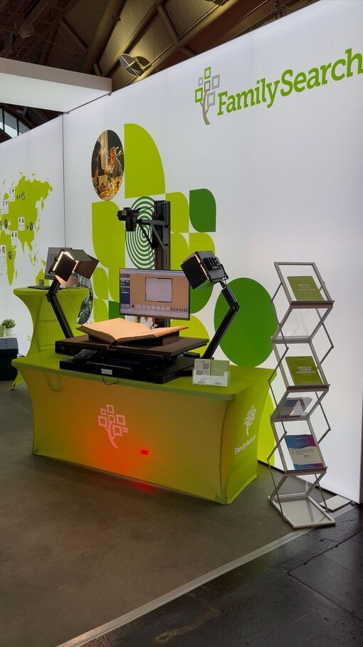 Digitalisierungsstation mit Buchscanner und Monitor, präsentiert bei einer Familienforschungsausstellung. Im Hintergrund Logowand.