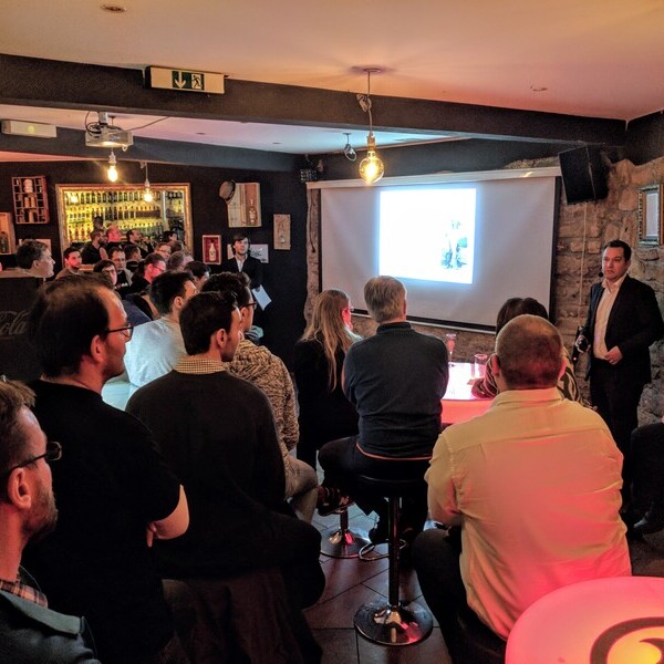 Personen verfolgen eine Präsentation in einem gemütlichen Barambiente. Ein Redner steht neben einer Leinwand.