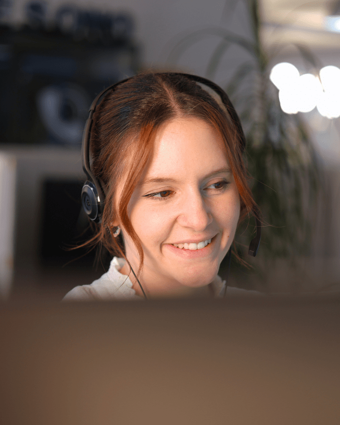 Frau mit Headset arbeitet an einem Computerarbeitsplatz in einem Büro, lächelnd und konzentriert auf den Bildschirm.