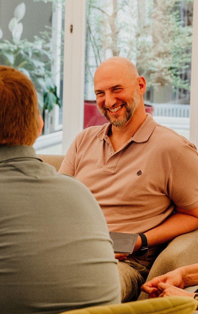 Unbekannte Person in einem Gespräch, lächelnd, sitzt in einem modernen, hellen Raum mit Pflanzen im Hintergrund.