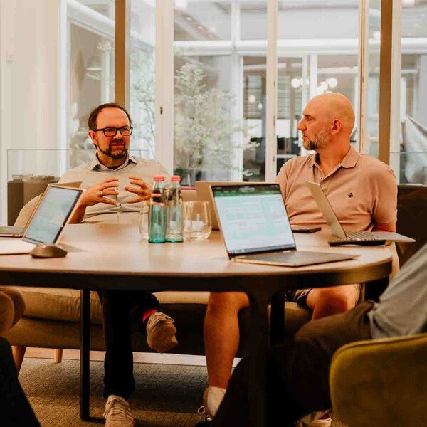 Vier Personen in einer Geschäftssitzung an einem runden Tisch mit Laptops und Notizen, informelle Atmosphäre.