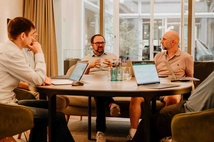 Vier Personen in einer Geschäftssitzung an einem runden Tisch mit Laptops und Notizen, informelle Atmosphäre.