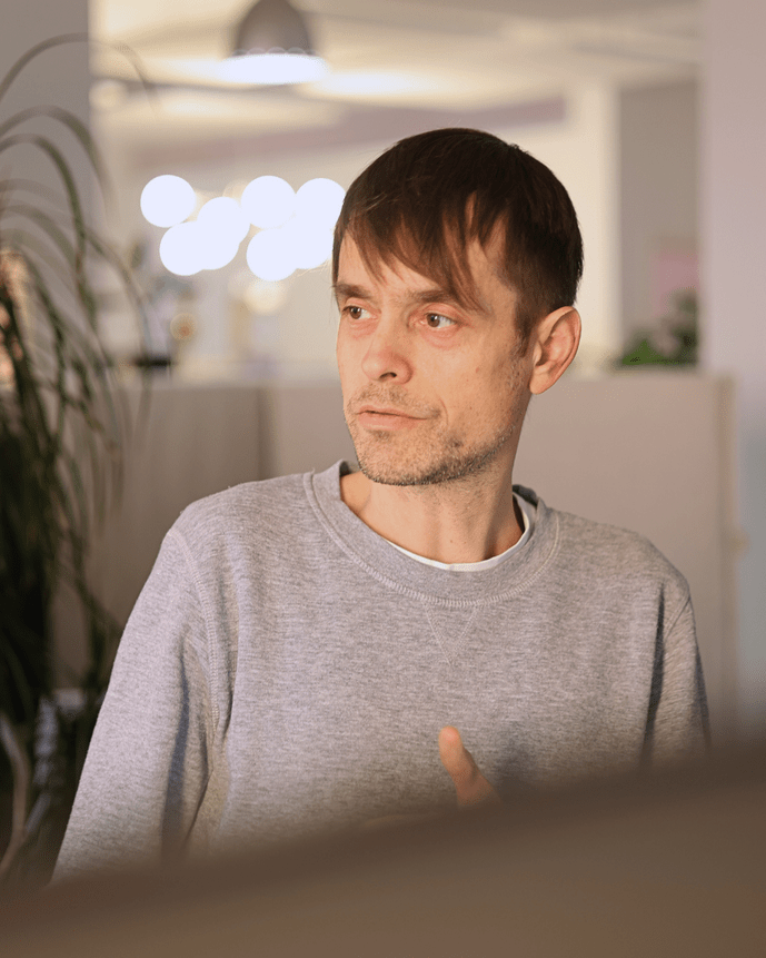 Eine Person in Freizeitkleidung sitzt in einem modernen Büro, im Hintergrund ist eine Pflanze und verschwommenes Licht zu sehen.