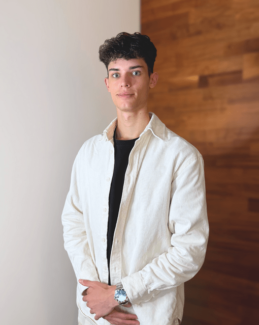 Eine Person in weißem Hemd steht vor einer hölzernen Wand und blickt in die Kamera.