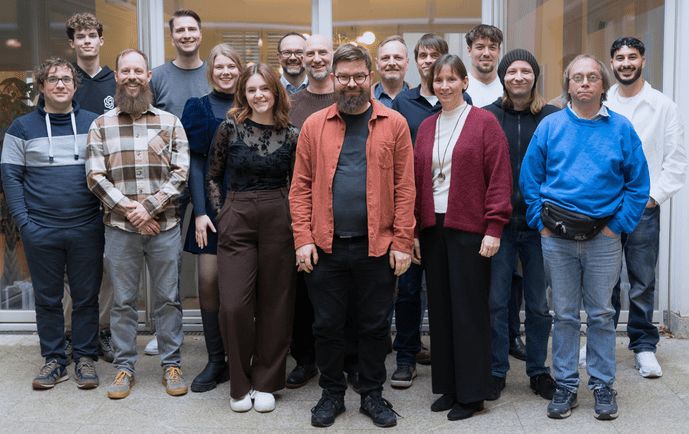 Eine Gruppe von 16 Menschen posiert in einem Flur vor einer Glaswand zu einem Gruppenfoto.
