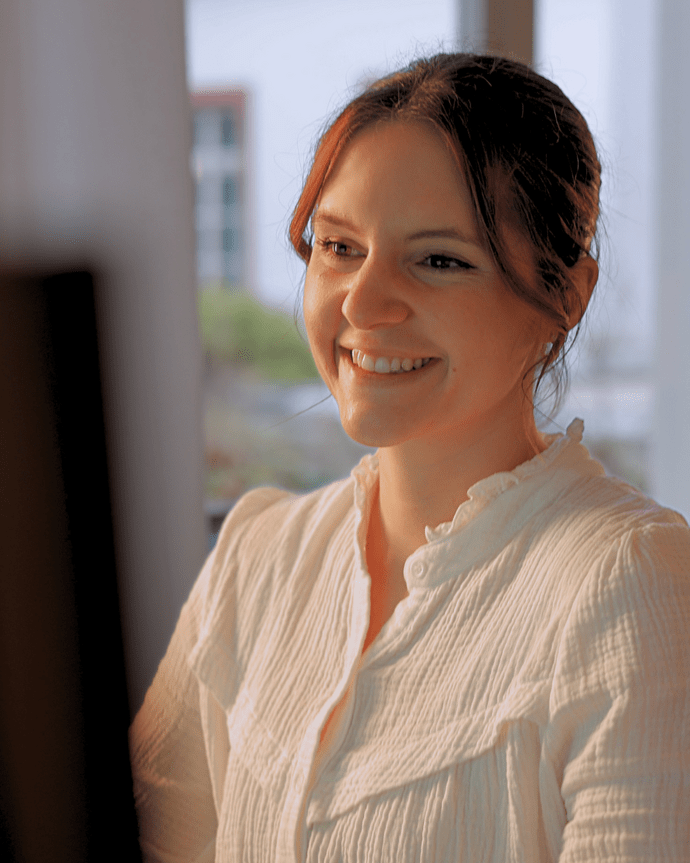 Eine Frau sitzt lächelnd vor einem Computer in einem hellen Raum mit Fenstern.