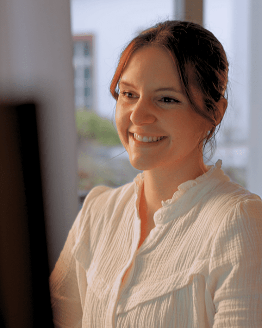 Eine Frau sitzt lächelnd vor einem Computer in einem hellen Raum mit Fenstern.