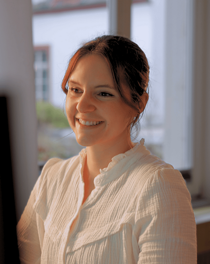 Eine Frau sitzt lächelnd vor einem Computer in einem hellen Raum mit Fenstern.