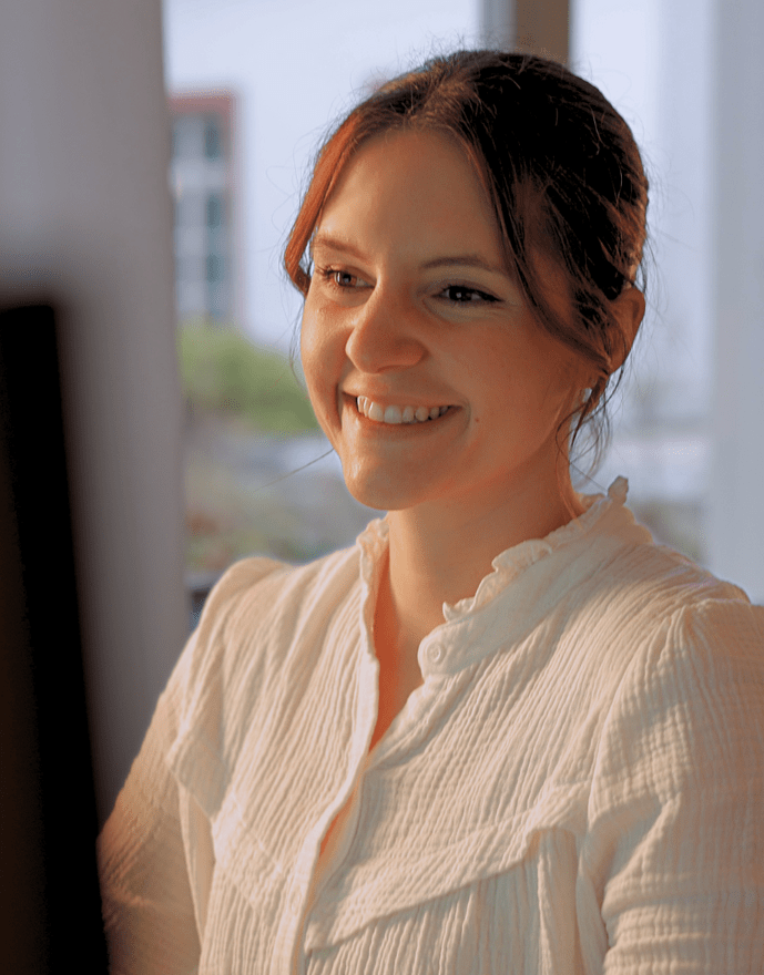 Eine Frau sitzt lächelnd vor einem Computer in einem hellen Raum mit Fenstern.