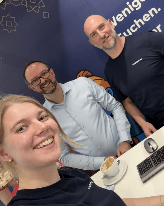 Drei Personen stehen lächelnd an einem Stand, eine junge Frau macht ein Selfie. Im Hintergrund ein dunkelblauer Banner.