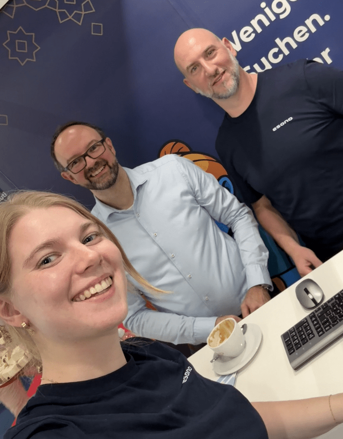 Drei Personen stehen lächelnd an einem Stand, eine junge Frau macht ein Selfie. Im Hintergrund ein dunkelblauer Banner.