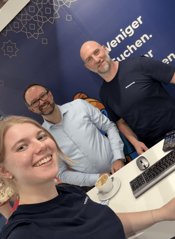 Drei Personen stehen lächelnd an einem Stand, eine junge Frau macht ein Selfie. Im Hintergrund ein dunkelblauer Banner.