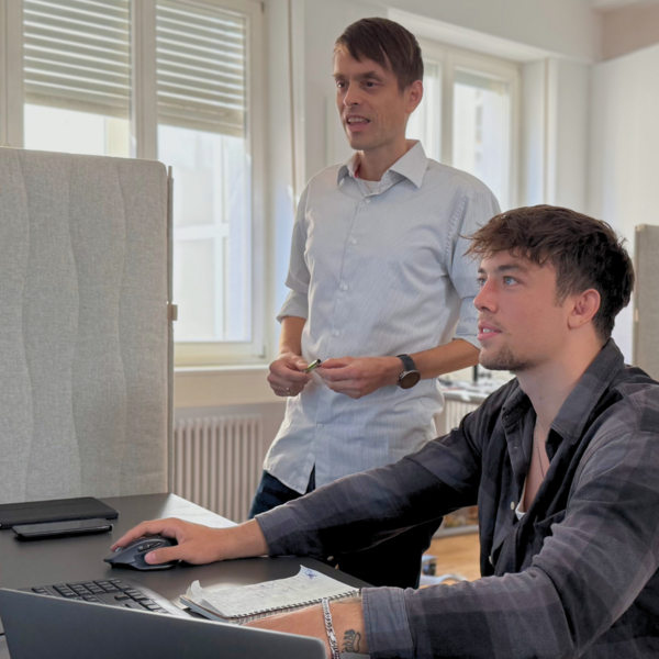 Zwei Männer arbeiten am Computer. Der sitzende Mann bedient Maus und Tastatur, der andere steht daneben und spricht.