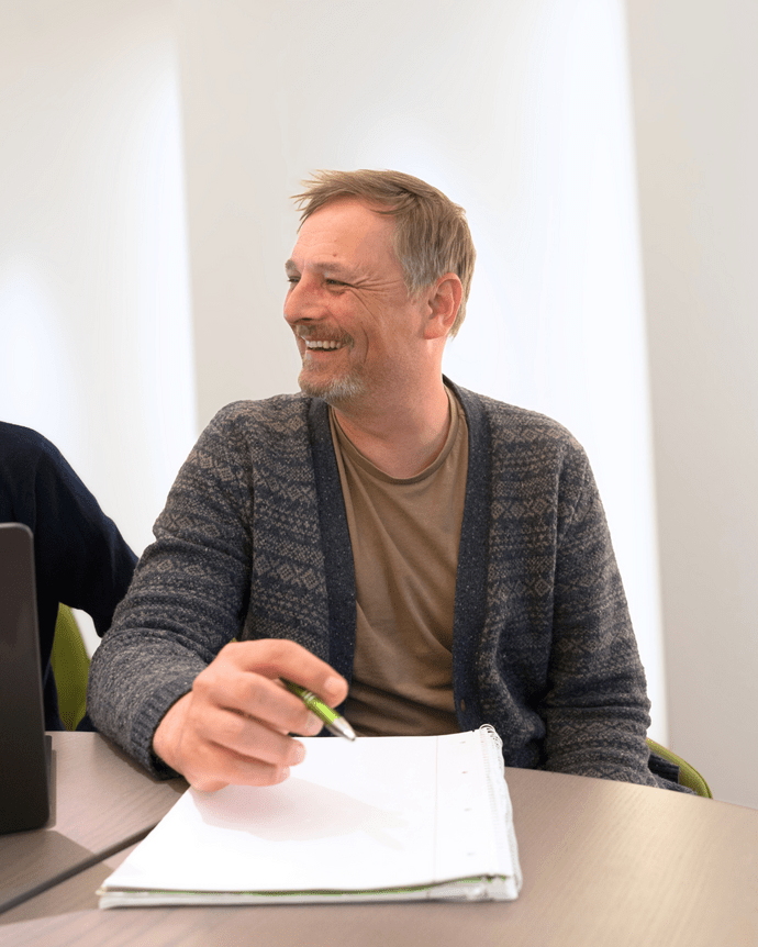 Mann lächelnd bei einem Treffen, Stift und Notizblock in der Hand, sitzt an einem Tisch.