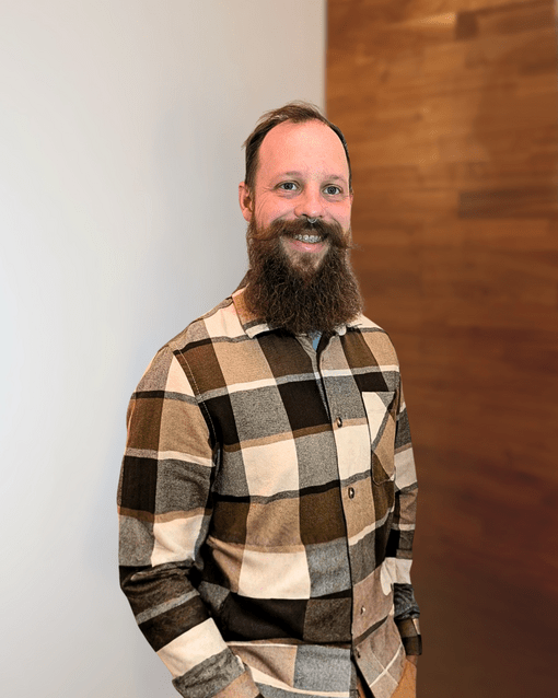 Ein Mann mit Vollbart und karierter Hemd steht vor einer teils weißen, teils holzvertäfelten Wand.
