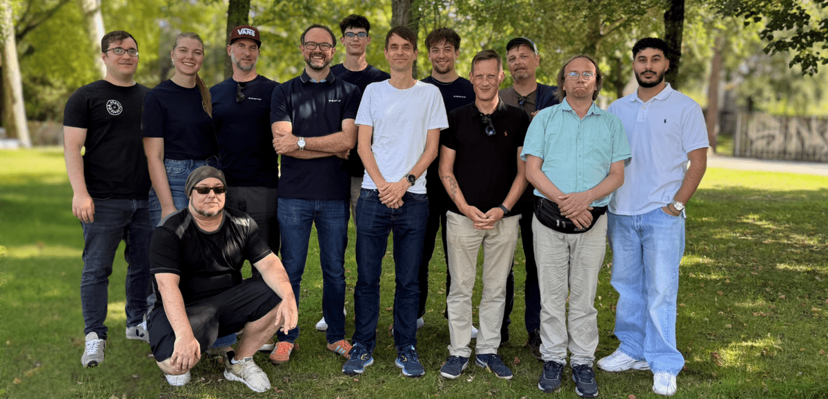 Gruppe von 13 Personen posiert im Freien vor Bäumen auf grasbewachsenem Gelände bei Tageslicht.