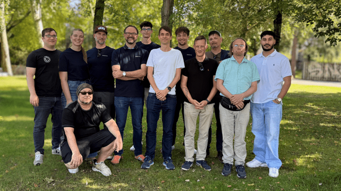 Gruppe von 13 Personen posiert im Freien vor Bäumen auf grasbewachsenem Gelände bei Tageslicht.