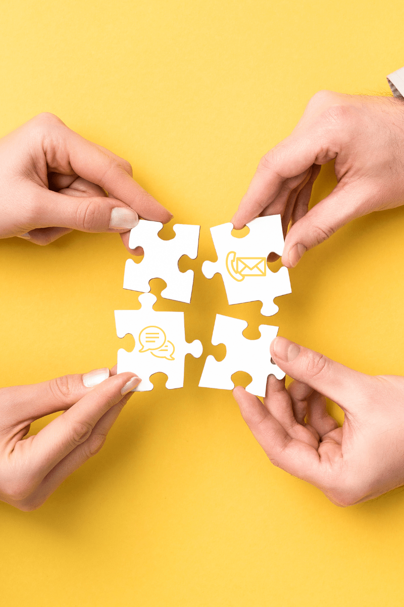 Vier Hände halten zusammenpassende Puzzleteile mit Kommunikationssymbolen vor gelbem Hintergrund.