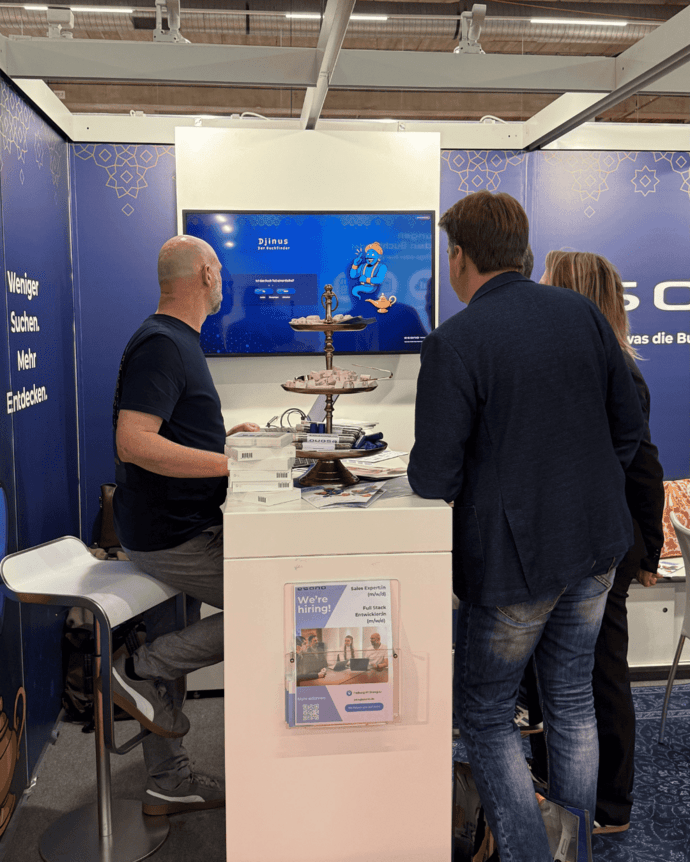 Drei Personen betrachten einen Bildschirm auf einer Messe. Ein Tisch mit Broschüren und eine Wand mit Text im Hintergrund.