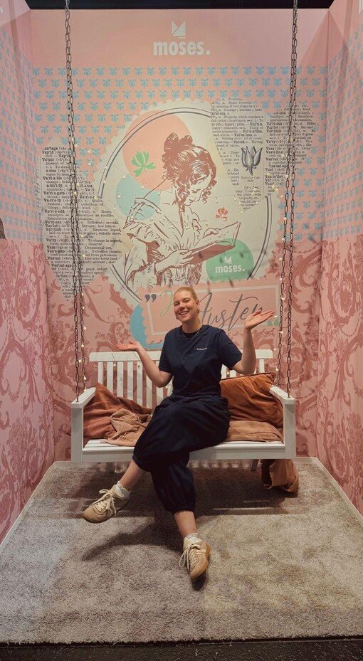 Person auf einer Schaukel; rosa Hintergrund mit grafischem Design, Schriftzug und Ketten als Schaukelaufhängung.