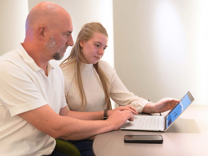 Zwei Personen arbeiten gemeinsam an einem Laptop an einem Tisch, auf dem auch ein Smartphone liegt.