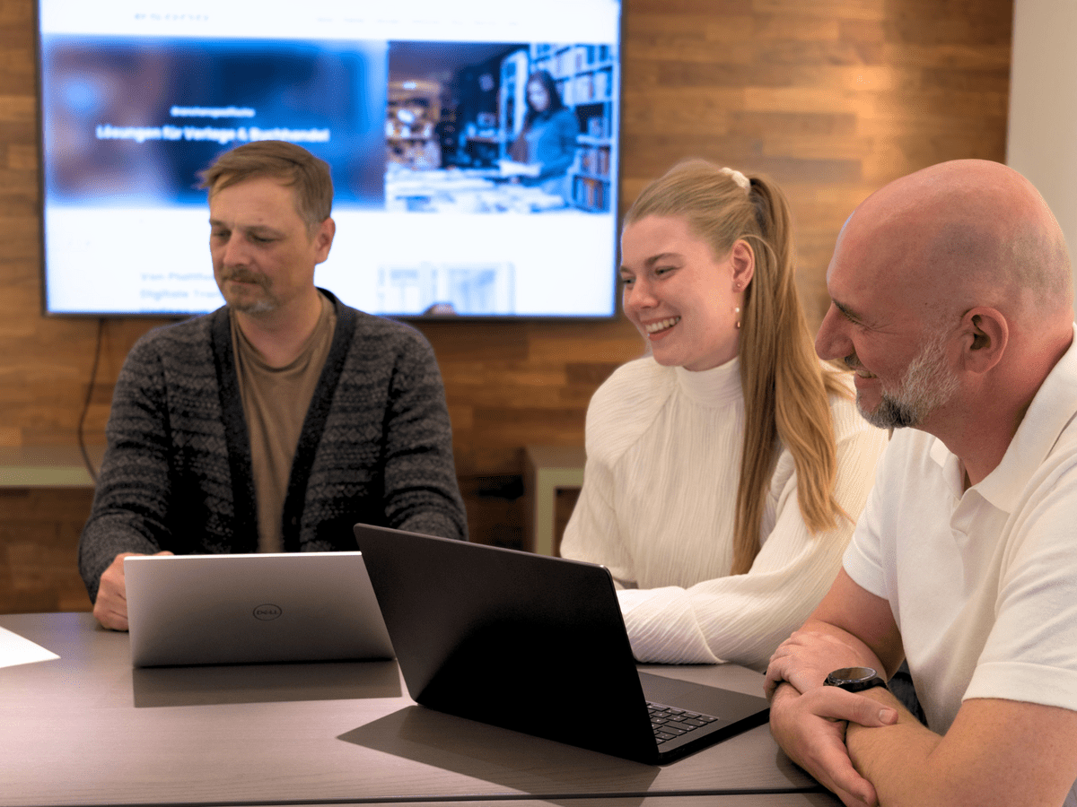 Drei Personen bei einer Besprechung am Tisch mit Laptops, im Hintergrund ein großer Bildschirm.