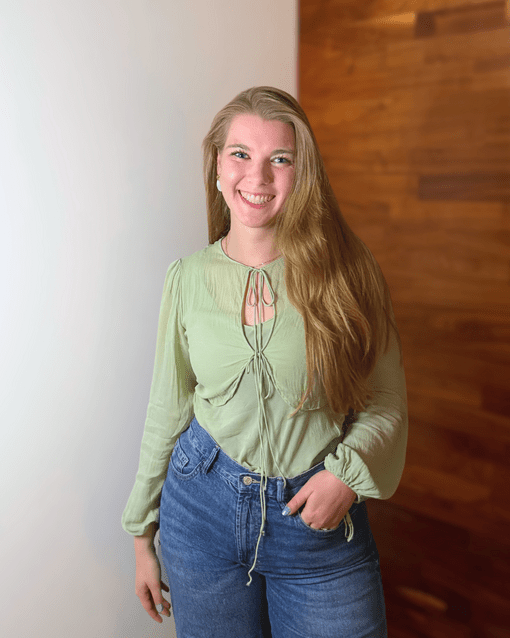 Junge Frau in grüner Bluse und blauer Jeans steht lächelnd vor holzvertäfeltem Hintergrund.