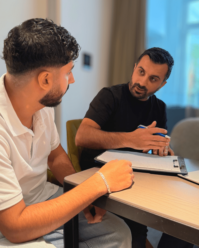 Zwei Männer diskutieren an einem Tisch. Einer hält ein Klemmbrett, der andere hört aufmerksam zu.