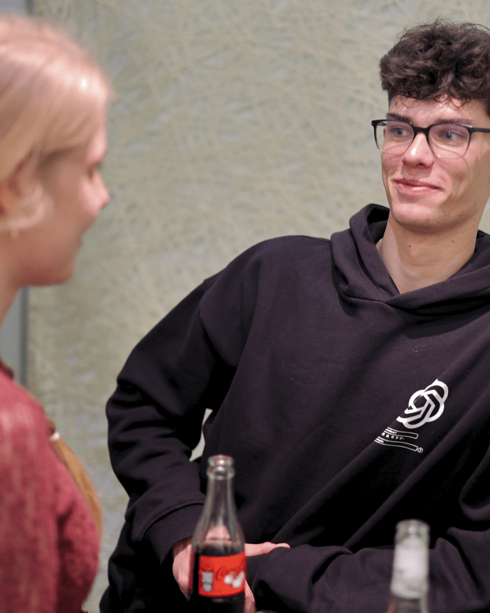 Zwei Personen im Gespräch an einem Tisch, leere Flaschen stehen vor ihnen. Eine trägt einen Hoodie, die andere hat blondes Haar.