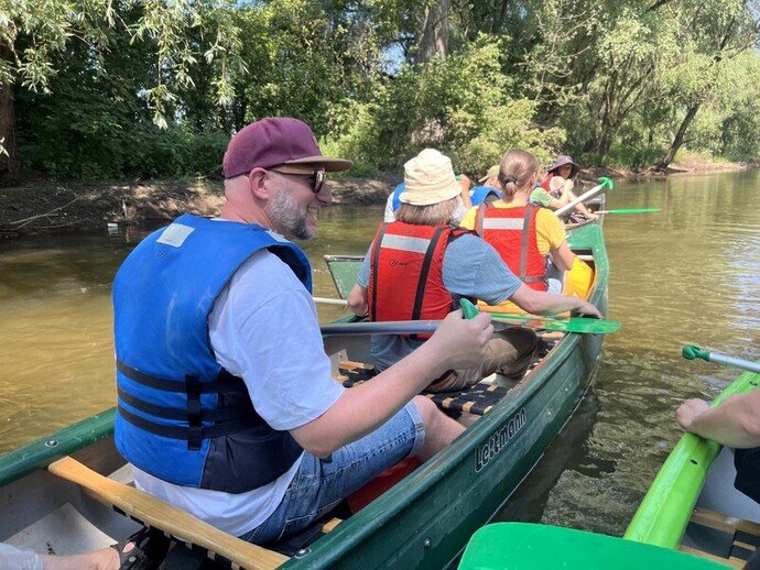 Menschen in Kanus auf einem ruhigen Fluss, tragen Rettungswesten und paddeln nahe einer bewaldeten Uferregion.
