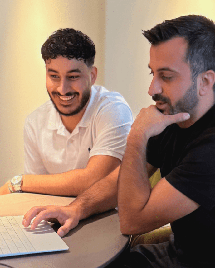 Zwei Männer sitzen an einem Tisch und schauen konzentriert auf einen Laptop. Ein Glas Wasser steht daneben.