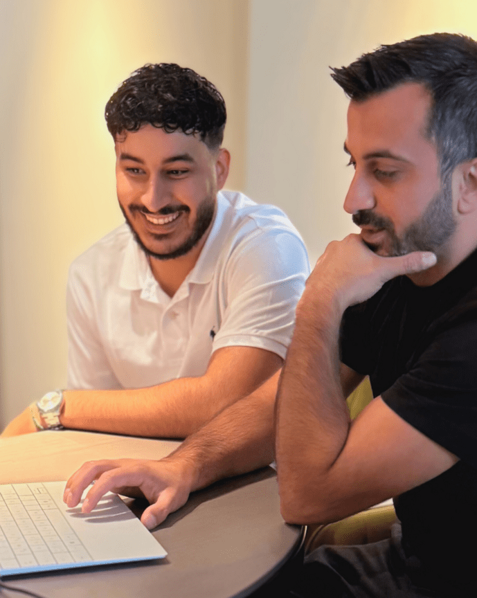 Zwei Männer sitzen an einem Tisch und schauen konzentriert auf einen Laptop. Ein Glas Wasser steht daneben.