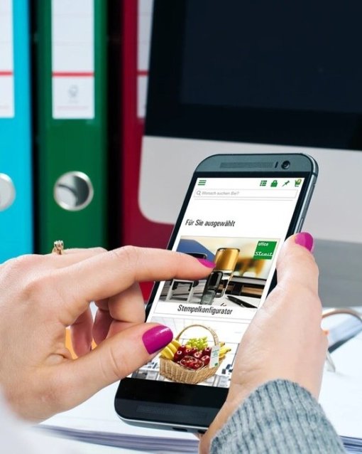 Person nutzt ein Smartphone vor einem Computerbildschirm, im Hintergrund sind bunte Ordner zu sehen.