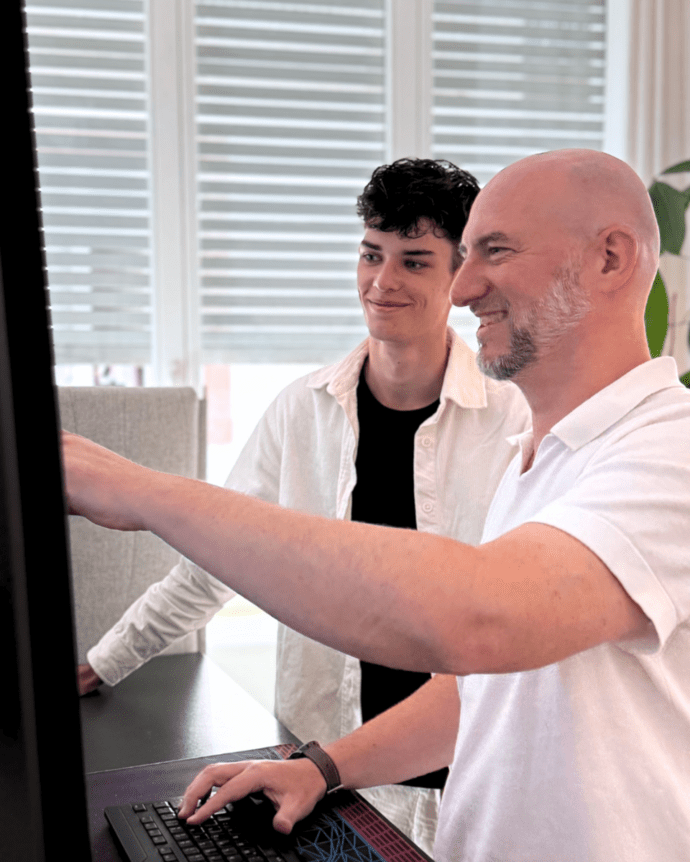 Zwei Männer arbeiten lächelnd an einem Computer in einem modernen Büro mit einer großen Pflanze im Hintergrund.