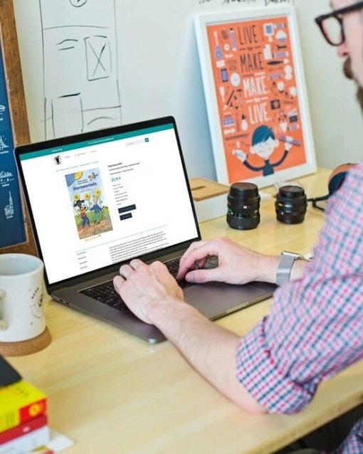 Person arbeitet am Laptop auf einem Schreibtisch mit Büchern, Pflanzen und Kameralinsen. An der Wand hängen Poster.