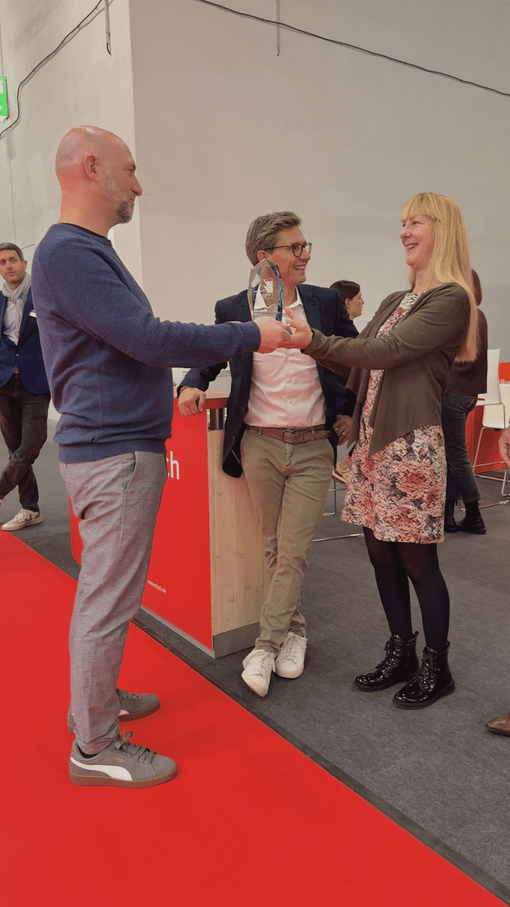 Drei Personen stehen auf rotem Teppich; eine Frau überreicht einem Mann einen Preis, während ein anderer Mann zusieht.