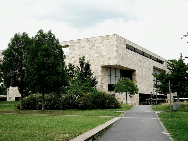 Moderne Bibliothek mit Steinfassade, umgeben von Bäumen und Grünflächen, angrenzend an einen Gehweg unter bewölktem Himmel.