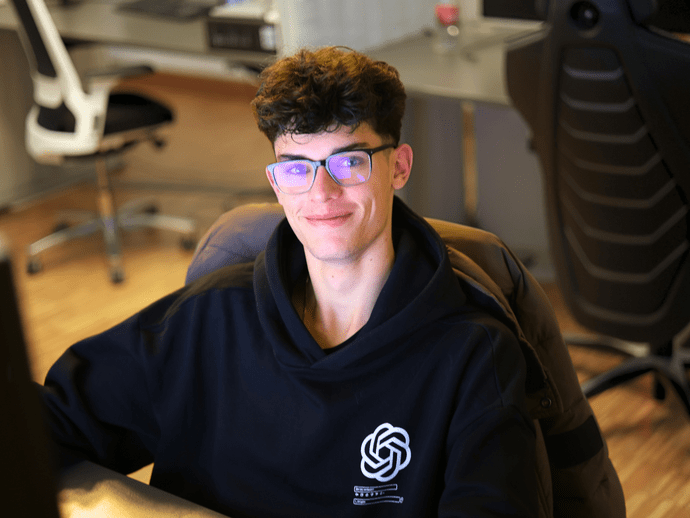 Ein lächelnder Mann mit Brille und schwarzem Hoodie sitzt an einem Schreibtisch in einem modernen Büro.
