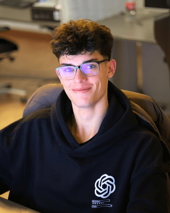 Ein lächelnder Mann mit Brille und schwarzem Hoodie sitzt an einem Schreibtisch in einem modernen Büro.