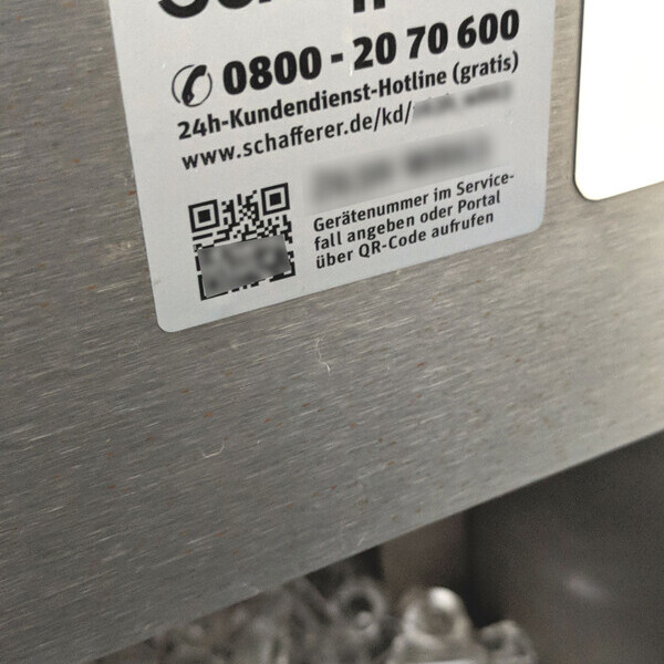 Silberne Metalloberfläche mit Aufkleber „Schafferer“, Hotline-Nummer und QR-Code, daneben transparente Plastikkappen.