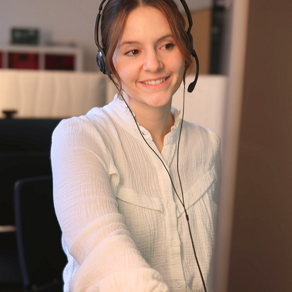 Eine Frau mit Headset lächelt und arbeitet an einem Computer, in einem Büro mit Regalen im Hintergrund.