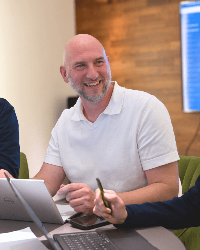 Ein Mann mit Glatze lächelt bei einem Treffen in einem modernen Büro, umgeben von Laptops und blauer Präsentationsfolie.