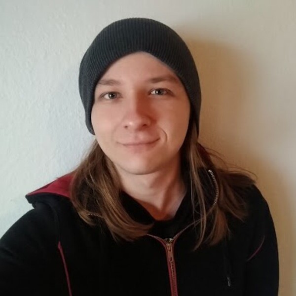 Eine Person mit langer Mütze und langen Haaren steht vor einer neutralen Wand und lächelt.