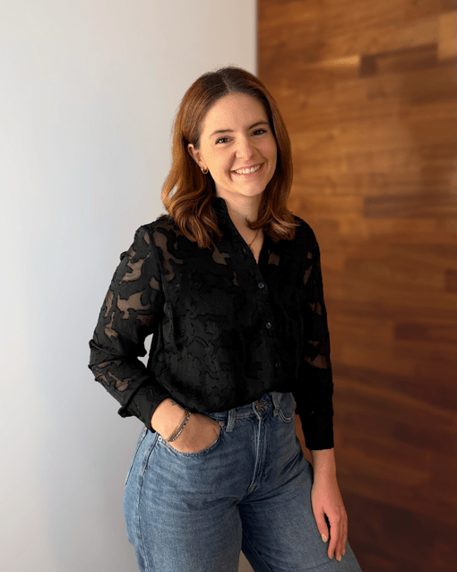 Eine Frau steht lächelnd in einem Raum mit Holzwand, trägt eine schwarze Bluse und blaue Jeans.