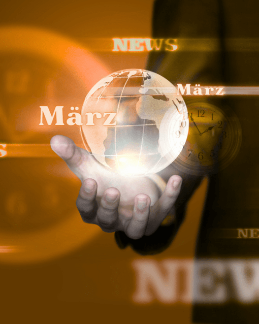 Hand hält rotierende Weltkugel mit Text "März" und "News" vor verschwommenem Uhr-Hintergrund in warmen Brauntönen.