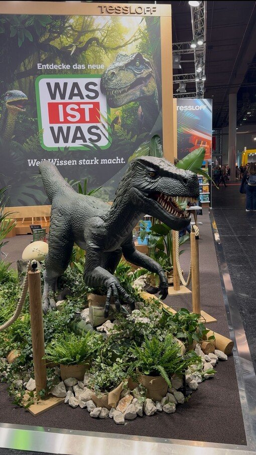 Dinosaurier-Modell in einer Messeausstellung vor einem Werbeplakat, umgeben von Pflanzen und Dekorationen.